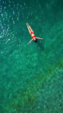 Kırmızı mayo giymiş bir kadının hava görüntüsü, kristal berrak deniz sularında rahatlama somutlaştırıcı bir şekilde sırtüstü uzanır. Yaz tatilleri. Dikey
