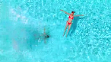 Çift havuzda rahatlar. Kadın sırtüstü yatarken, bir erkeğin havuza dalıp yüzdüğü tepeden aşağı manzara..