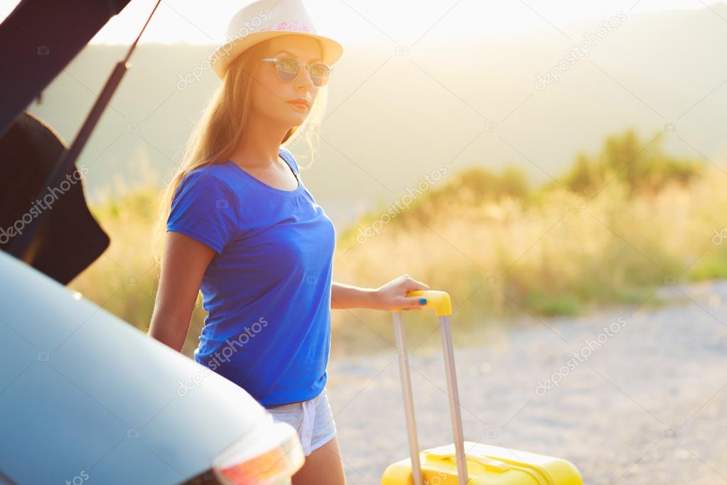 Woman with a yellow suitcase standing near the trunk of a car pa