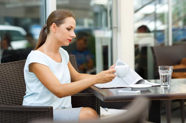 Mujer joven leyendo un periódico y bebiendo café de la mañana en un —  Fotos de Stock