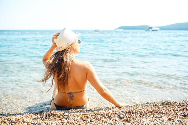 Genç kadın bir şapka ve bikini oturma plaj - üzerinden görüntülemek — Stok fotoğraf