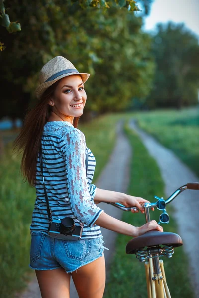 Όμορφη νεαρή γυναίκα με ένα καπέλο με ένα ποδήλατο σε ένα πάρκο — Φωτογραφία Αρχείου