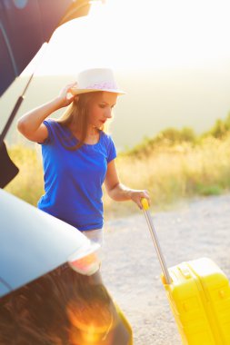 Young woman with a yellow suitcase standing near the trunk of a 