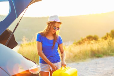 Young woman with a yellow suitcase standing near the trunk of a 