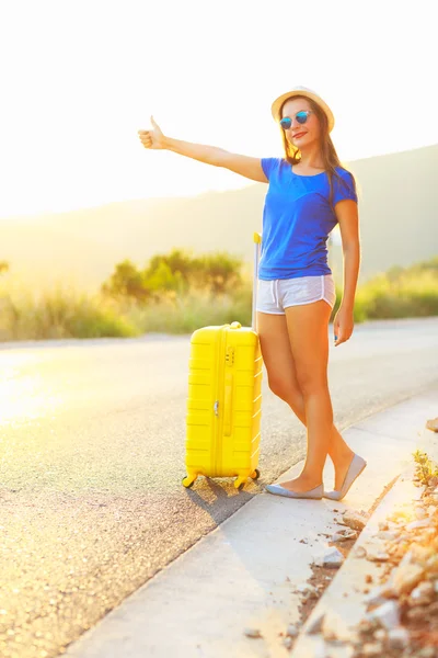 Молодая женщина с большим пальцем и желтым чемоданом едет дальше — стоковое фото