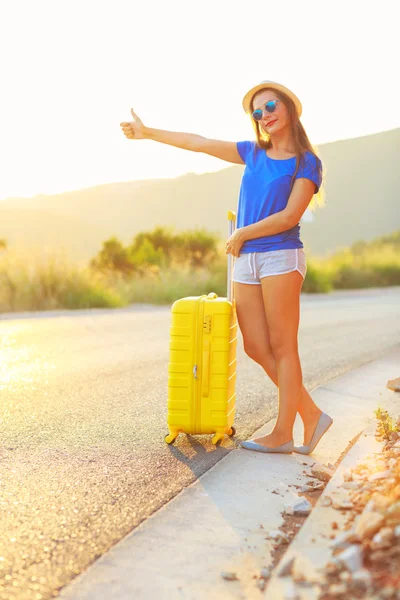 Mujer joven con una maleta amarilla está viajando en el camino hitc —  Fotos de Stock