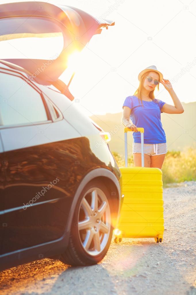 Woman with a yellow suitcase standing near the trunk of a car pa