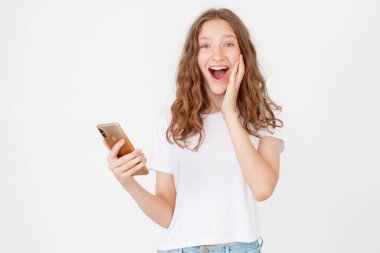 Şaşkın genç kız cep telefonunu olumlu bir ifadeyle kullanıyor, geniş bir şekilde gülümsüyor, günlük kıyafetler giyiyor ve beyaz arka planda tek başına duruyor. İnsanların yaşam tarzı konsepti. Happy Teen Cep telefonu kullanarak SMS mesajı yazma Winer