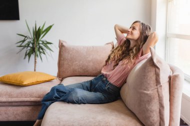 Huzurlu genç bayan oturma odasındaki kanepede dinleniyor. Sakin, mutlu kadın serbest çalışan, ellerini başının arkasına koyuyor, gözlerini kapıyor ve evde uzak çalışma gününde ara veriyor.