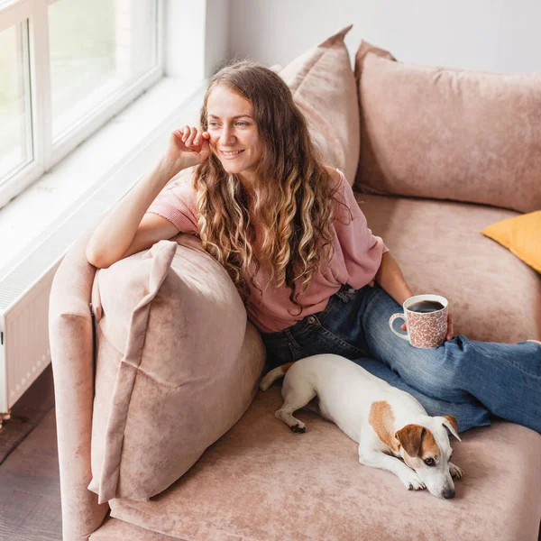 静かで穏やかな中年の女性は 自宅でソファの上に熱いお茶のカップで休憩を取ります 内容魅力的な幸せな女性楽しんでコーヒーと白昼夢上のソファとともに犬 — ストック写真