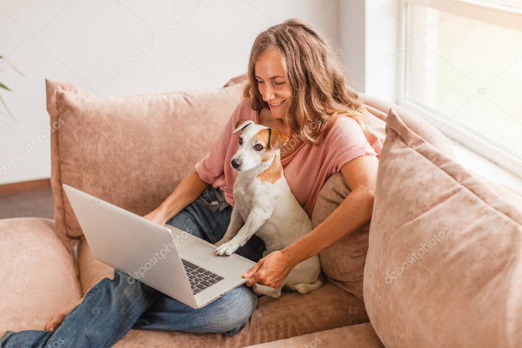Happy girl with dog sitting on sofa. Beautiful cheerful woman using silver laptop while sitting on sofa in living room at home