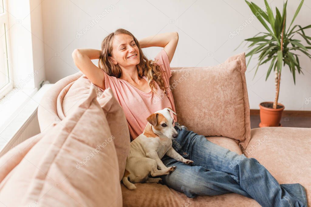 Serene young woman pretty relaxing on couch with her dog in living room. Calm happy female freelancer crossing hands behind head, taking a break during remote work day at home