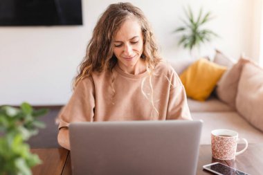 Genç neşeli, mutlu, pozitif, güzel iş kadını evde oturup dizüstü bilgisayar kullanıyor.