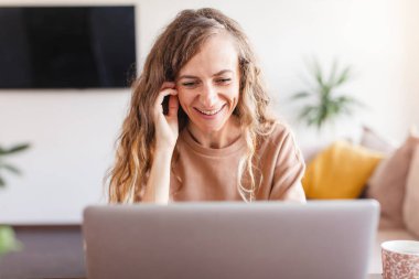 Evde dizüstü bilgisayarla çalışan mutlu, sıradan, güzel bir kadın. Bilgisayara bakan, gülümseyen, serbest çalışan bir kadın.