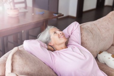 Evdeki rahat koltukta oturan sağlıklı orta yaşlı kadının portresi. Gülümseyen, 50 'li yaşlarda beyaz saçlı bir anne kameraya bakıyor, modern oturma odasında poz veriyor.