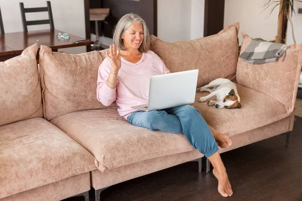 ソファに座っている間 熟女笑顔の女性がノートパソコンに手を振っています 女性言うことこんにちはでAビデオ会議チャットハッピーグレー毛の中年女性で家 — ストック写真