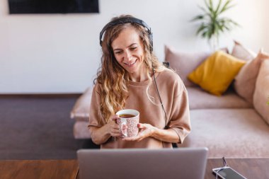 Kadın bir dizüstü bilgisayarda görüntülü sohbet okuyor. Mutlu kadın serbest çalışan uzak bir evde çalışıyor. Kulaklıklı kız görüntülü arama ile iletişim kuruyor.