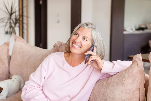 魅力的な成熟した灰色の髪の女性のソファに座って 通信を呼び出す 幸せな中年女性座っています上のソファに家と電話 — ストック写真