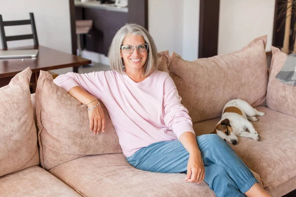 自宅で快適なソファに座って肖像幸せな健康的な中年の女性 快適な50代の白髪のお母さんを笑顔カメラを見て 現代のリビングルームでポーズ — ストック写真