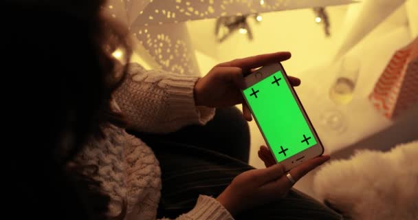 Mujer morena mirando la pantalla verde del teléfono inteligente sentado cerca del árbol de Navidad. — Vídeo de stock