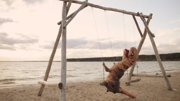 Enorma uppblåsbara dinosaurien svänger på en gunga på stranden vid solnedgången. — Stockvideo
