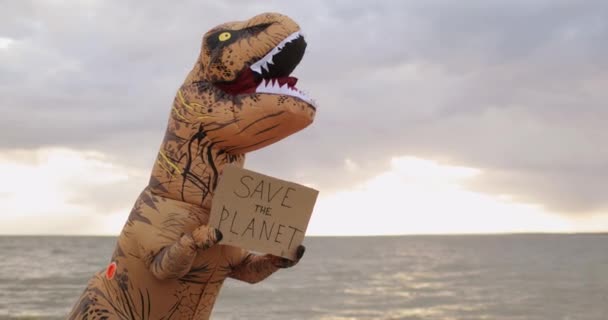Ogromny tyranozaur Rex dinozaur trzymający sztandar uratować planetę w łapach na plaży. — Wideo stockowe