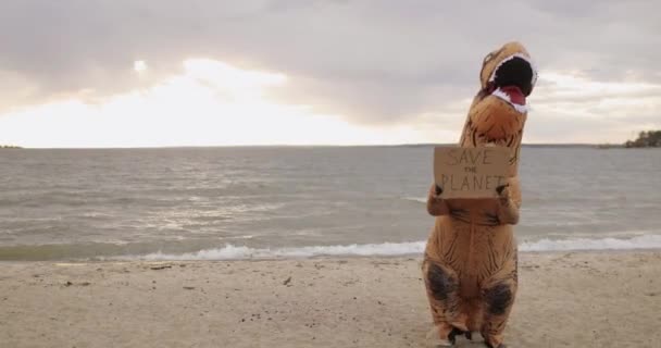 Huge dinosaur holds banner save the planet in paws, shows it to camera on beach. — Stock Video