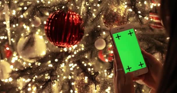 Mujer joven está viendo en la pantalla verde del teléfono inteligente sentado cerca del árbol de Navidad. — Vídeo de stock