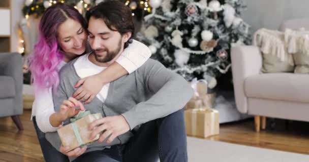 Молода жінка дарує подарунок своєму хлопцеві в новорічному інтер'єрі вдома . — стокове відео