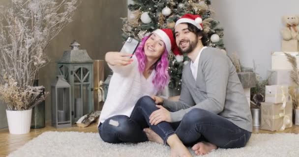 Jeune couple en chapeaux Santas fait selfie sur smartphone près de l'arbre de Noël. — Video