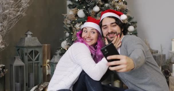 Man och kvinna i tomte hattar gör selfie på smartphone nära julgran. — Stockvideo