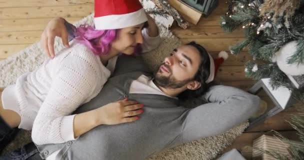 Verliebtes junges Paar in roten Hüten, das neben dem Weihnachtsbaum liegt und sich umarmt und küsst. — Stockvideo