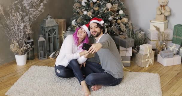 Fru och make i tomte hattar gör selfie på smartphone nära julgran. — Stockvideo