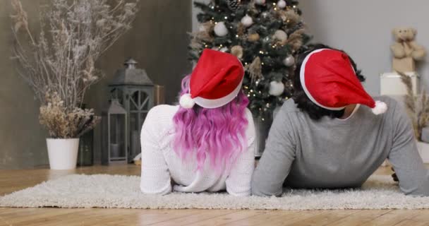 Hombre y mujer en los sombreros rojos de Santas acostados juntos cerca del árbol de Navidad, vista trasera. — Vídeo de stock