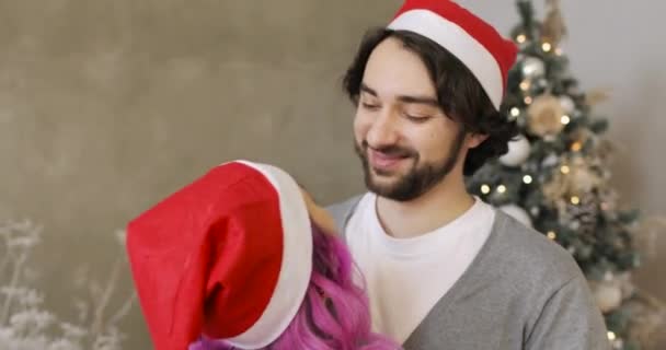 Joven esposo y esposa en Santas sombreros rojos acurrucados cerca del árbol de Navidad en casa. — Vídeo de stock