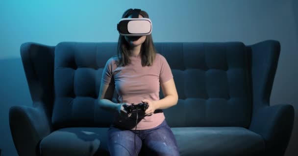 Mujer se sienta en el sofá en gafas de realidad virtual juega videojuego utilizando gamepad. — Vídeos de Stock