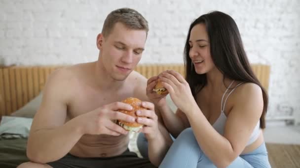 Jeune couple homme et femme mangent des hamburgers ensemble après l'entraînement à la maison. — Video