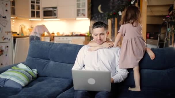 Far forsøger at arbejde på bærbar computer med to børn krammer ham i stuen. – Stock-video
