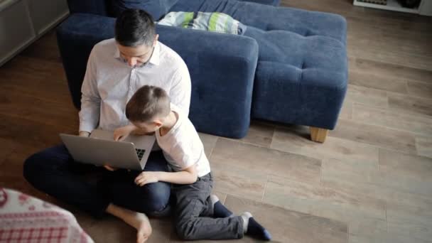爸爸正在教他的小儿子在家里的地板上用笔记本电脑工作. — 图库视频影像
