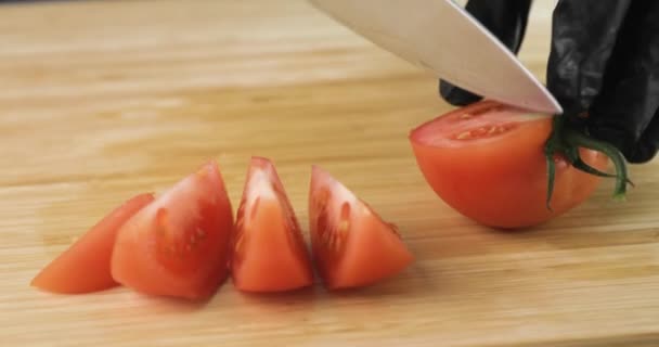 Кухарі руками в чорних рукавичках нарізають свіжий помідор на шматки на дерев'яній дошці . — стокове відео