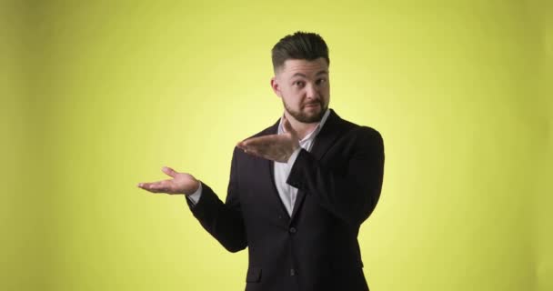 Mann im Büroanzug zeigt Zeigefinger zur Seite auf leere Handfläche auf gelbem Hintergrund. — Stockvideo