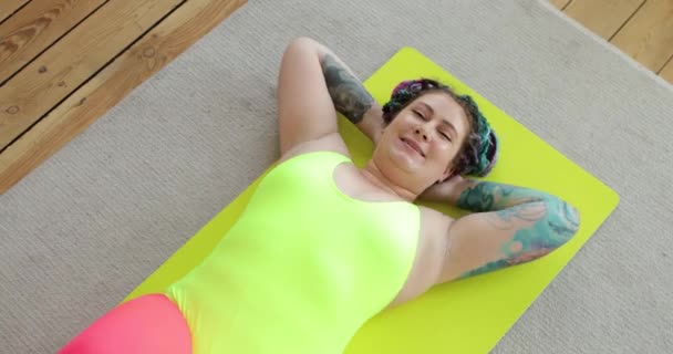 Толстая женщина новичок в спорте делает абс упражнения дома лежа на коврике, вид сверху. — стоковое видео