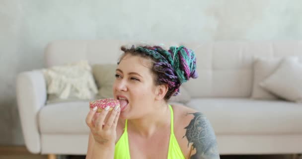 Fat woman in sportswear sitting on mat enjoying eating her doughnut at home. — Stock Video