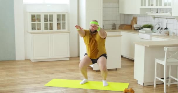 Skægget tyk svedig mand i gul våd sportstøj gør squats motion derhjemme. – Stock-video