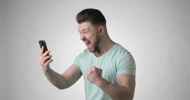 Hombre joven en camiseta verde mirando en el teléfono inteligente gritando haciendo gesto ganador. — Vídeo de stock
