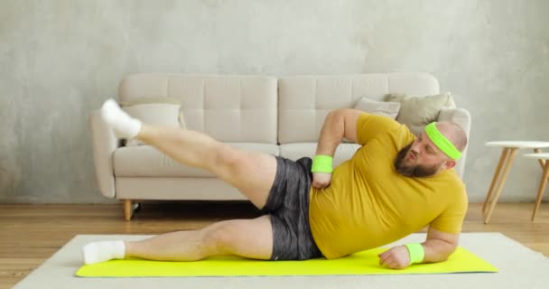 Gordo no sportswear amarelo fazendo exercícios de levantamento de pernas deitado no tapete em casa. — Vídeo de Stock