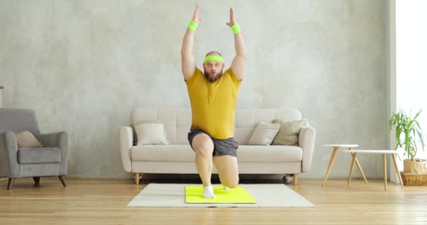 Tyk mand gør squats i lunger motion med hænderne oppe stående på måtten derhjemme. – Stock-video