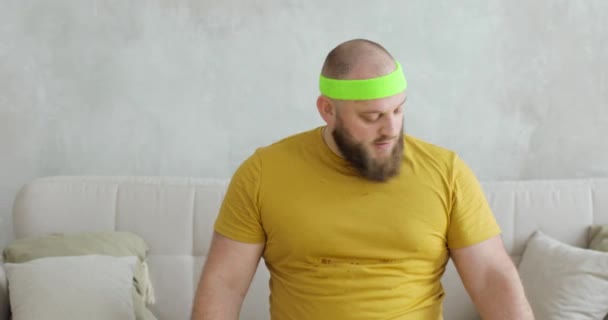 Vtipný tlustý muž ve sportovním oblečení zvedání činky rukama sedí na pohovce doma. — Stock video