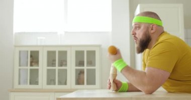 Sebze yiyen şişman bir adam masada oturmuş dambılla el egzersizi yapıyor..