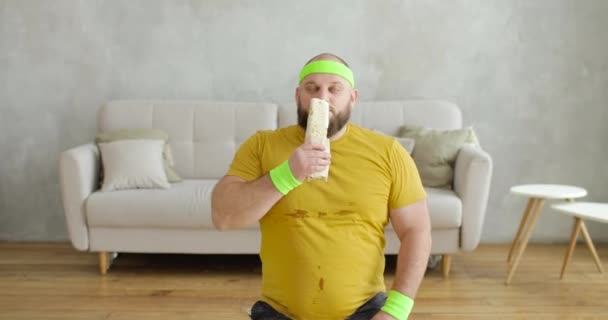 Grasso uomo sta mangiando shawarma seduto sul tappeto fitness a casa dopo gli esercizi sportivi. — Video Stock